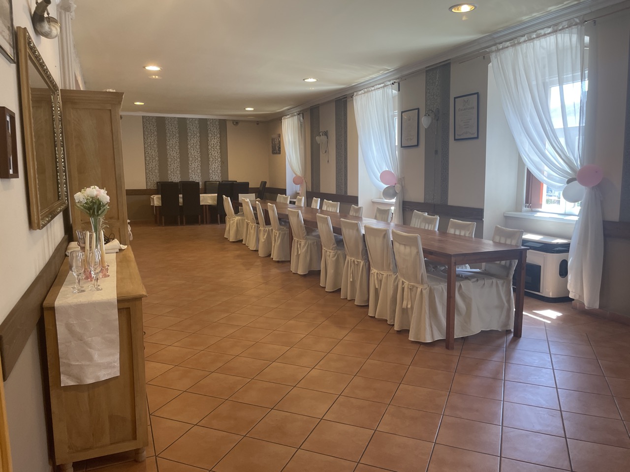 Tágas, világos rendezvényterem hosszú asztalokkal és elegáns székhuzatokkal