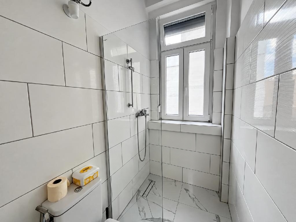 Zuhanyzó üveg zuhanyfallal, ablakkal és márványhatású padlóval a felújított fürdőszobában