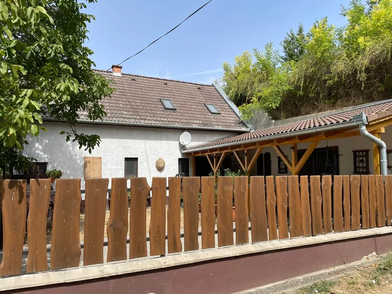 A ház cseréptetője, tetőablakai és fedett kiülője az udvari fa kerítés mögött.