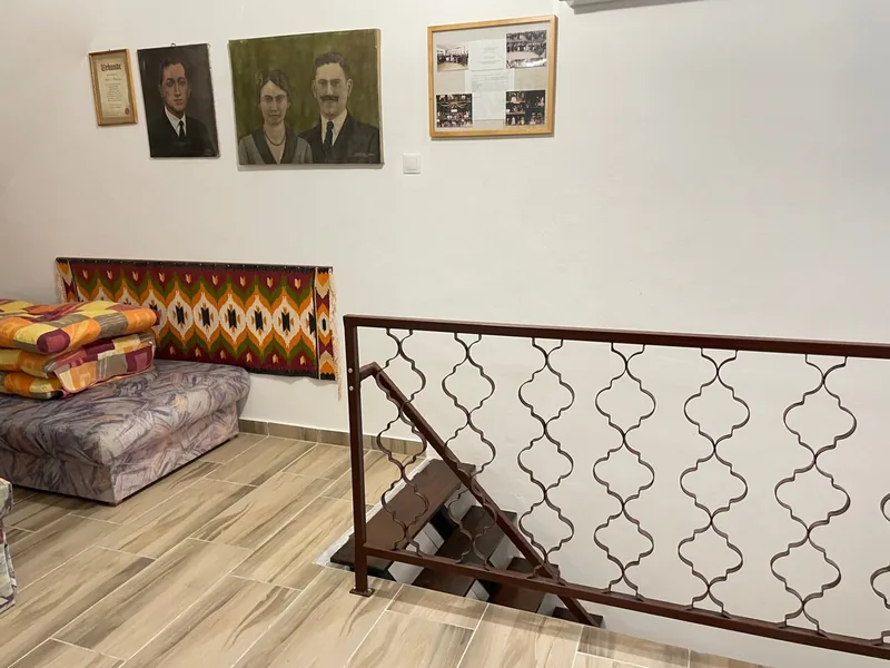 Fém lépcsőkorlát, mintás ülőbútorok és bekeretezett képek a világos falú belső térben.