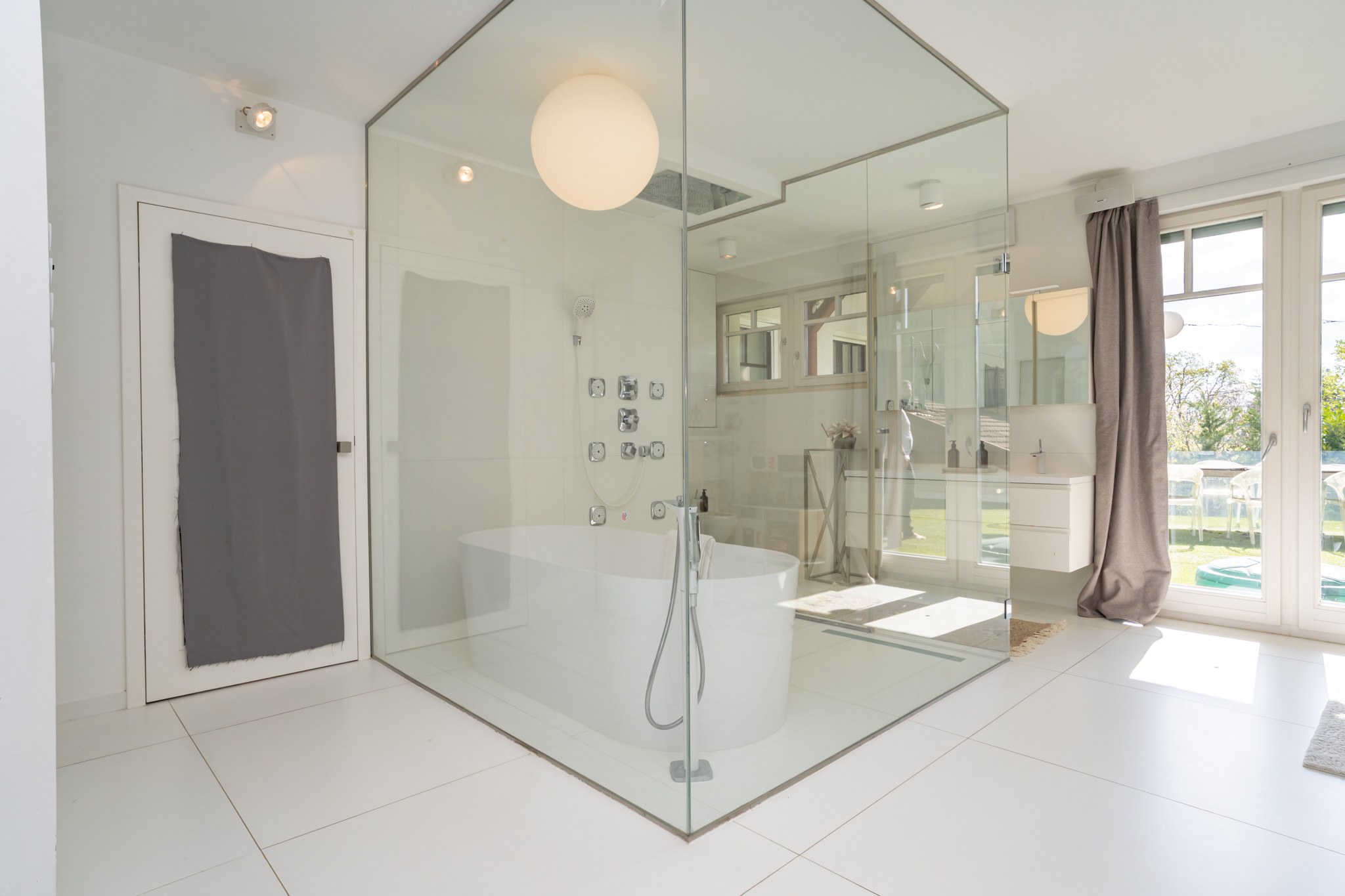A bright, modern bathroom with a freestanding bathtub and glass shower enclosure. Large windows look out onto the manicured garden.
