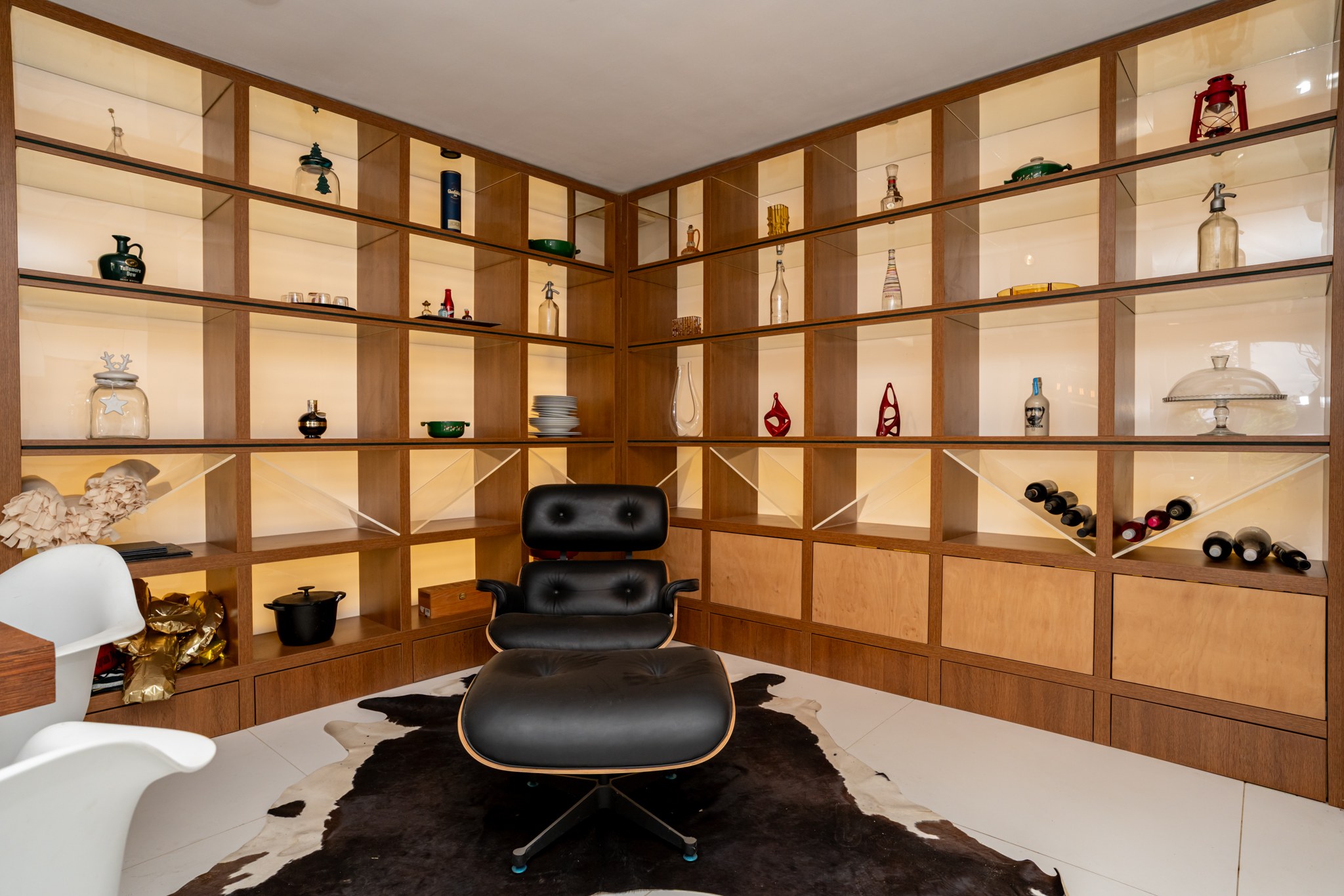 A modern-style built-in wooden shelving corner filled with decorative objects and wine bottles. An elegant black leather armchair takes centre stage on a fur rug. The atmosphere is modern and well-ordered.