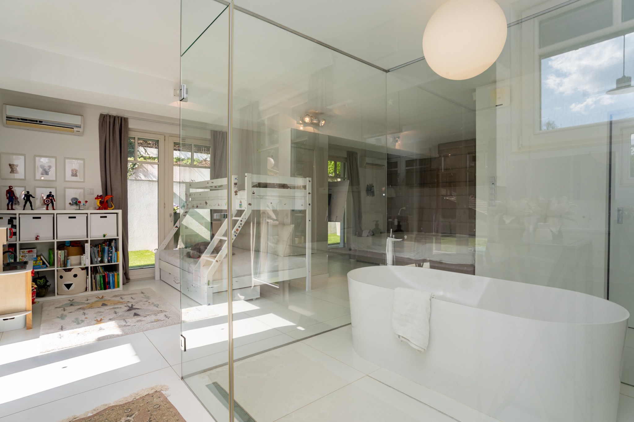 A modern interior where a bright children's room connects to a bathroom through a glass wall. The bathtub has a clean, contemporary design; the children's room features a bunk bed and numerous toys. Large windows and a glass door allow plenty of natural light and views of the garden.