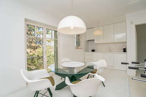 A modern, bright open-plan kitchen-dining room with minimalist white cabinetry and a stylish dining set. A large window looks out onto the green trees.
