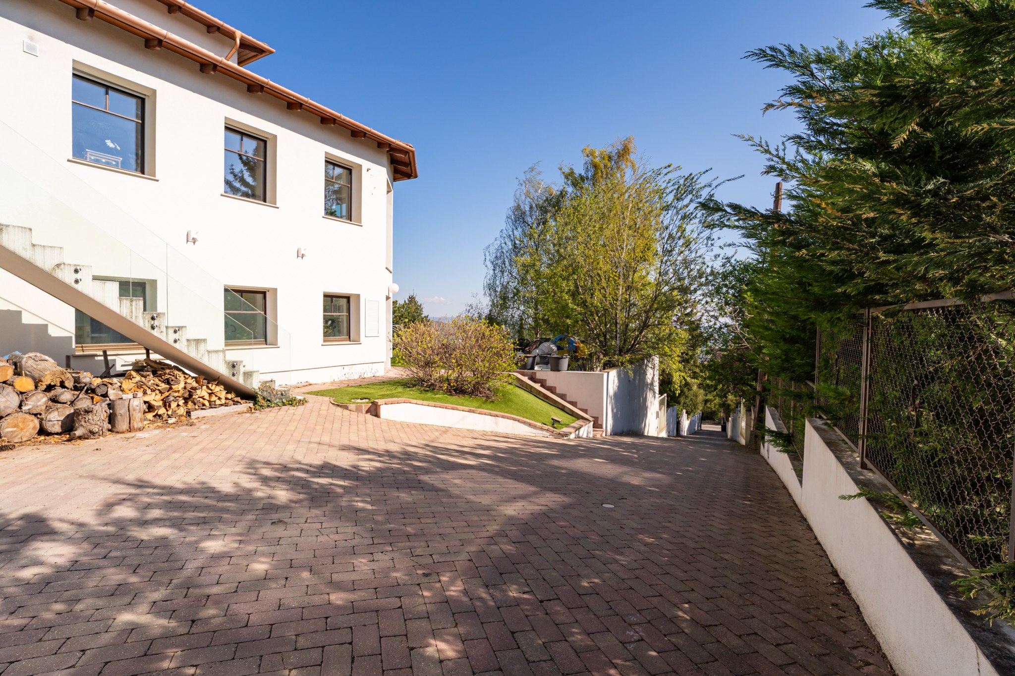 The photo shows the exterior of a white, modern family home with its paved parking area in sunny conditions. A external staircase with a glass balustrade is visible, along with a large stack of firewood beside the house.