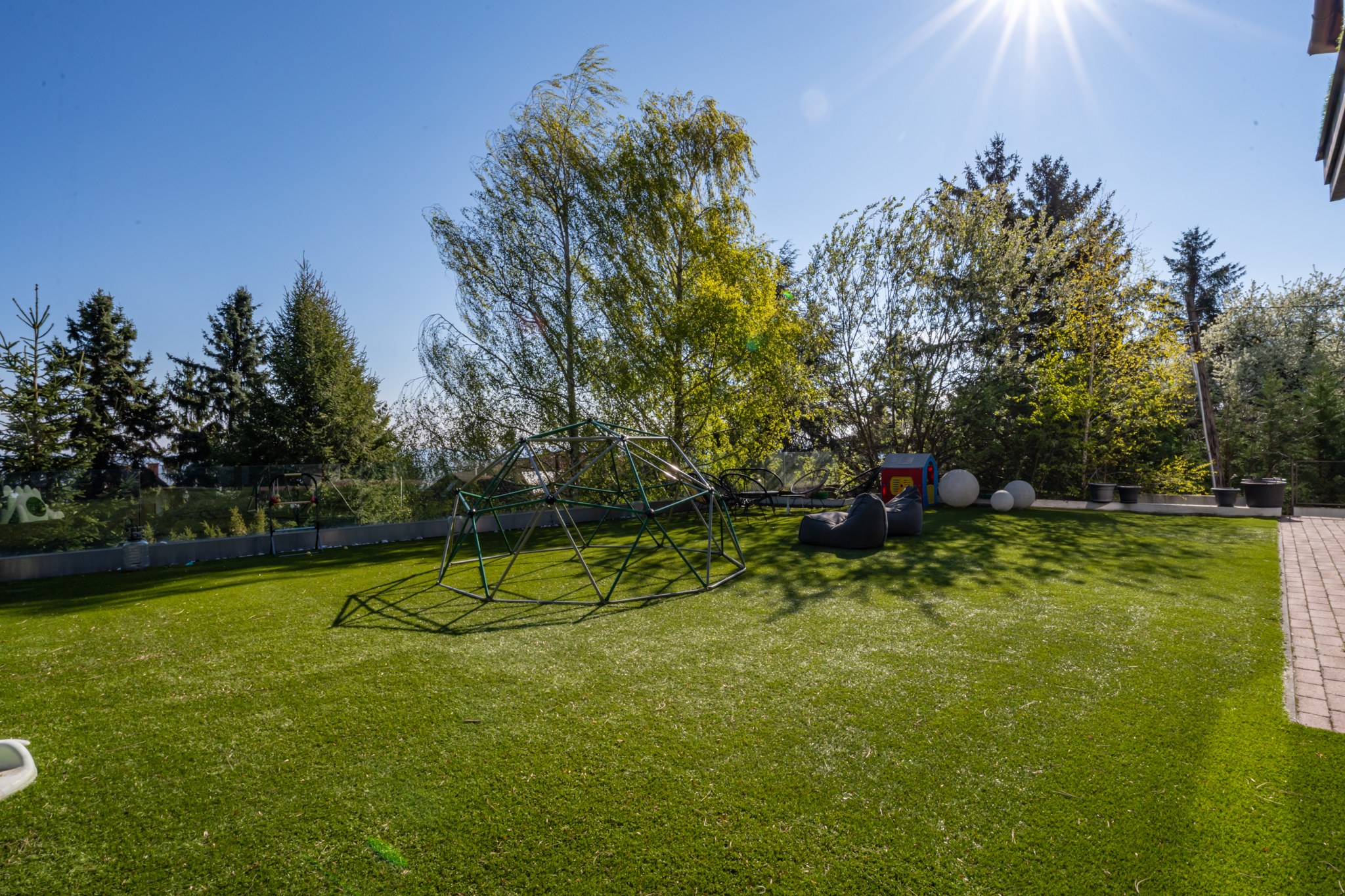 The photo shows a sun-filled, spacious outdoor space with artificial grass, children's play equipment and trees in the background. It conveys the feel of a modern, well-kept play courtyard.