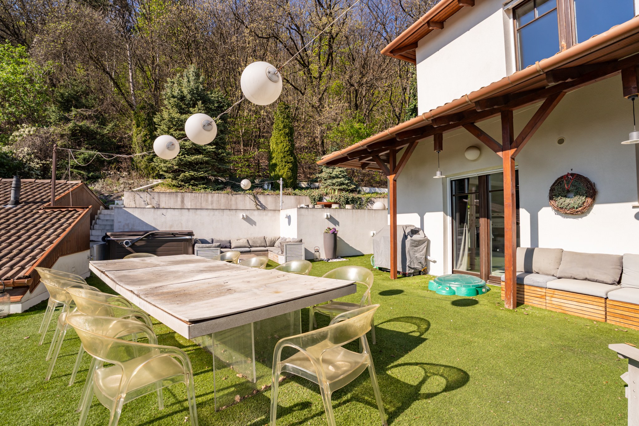 A spacious, modern terrace covered with artificial grass, featuring a dining area, a relaxation corner and a hot tub. The terrace offers views of the green, wooded hillside.