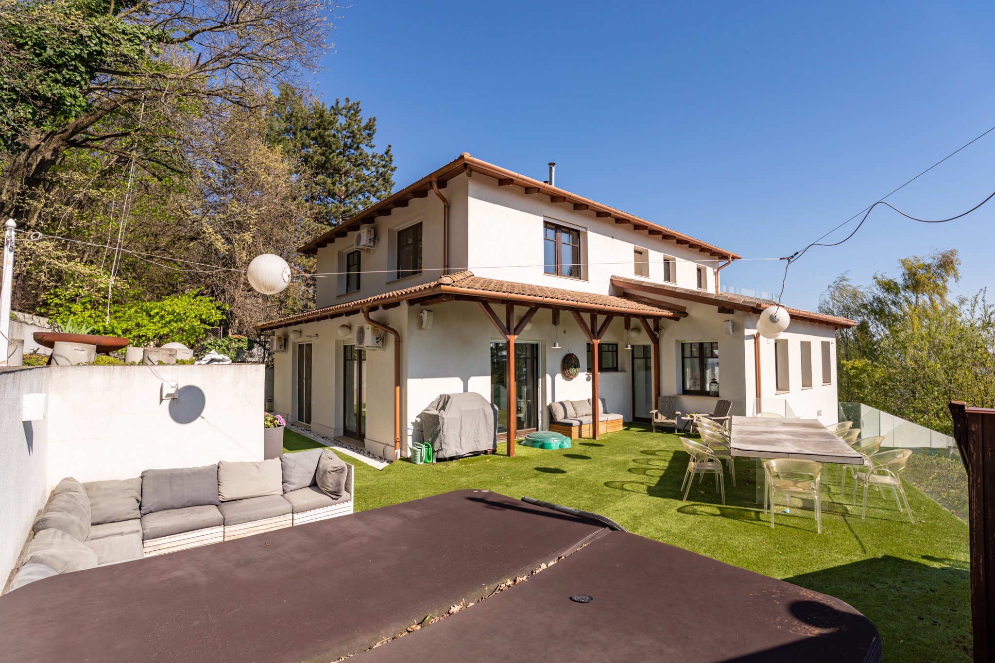 The rear garden of a modern white-walled family home in a terraced layout. The garden features artificial grass, a hot tub, an outdoor dining set and a comfortable seating area, surrounded by lush planting.