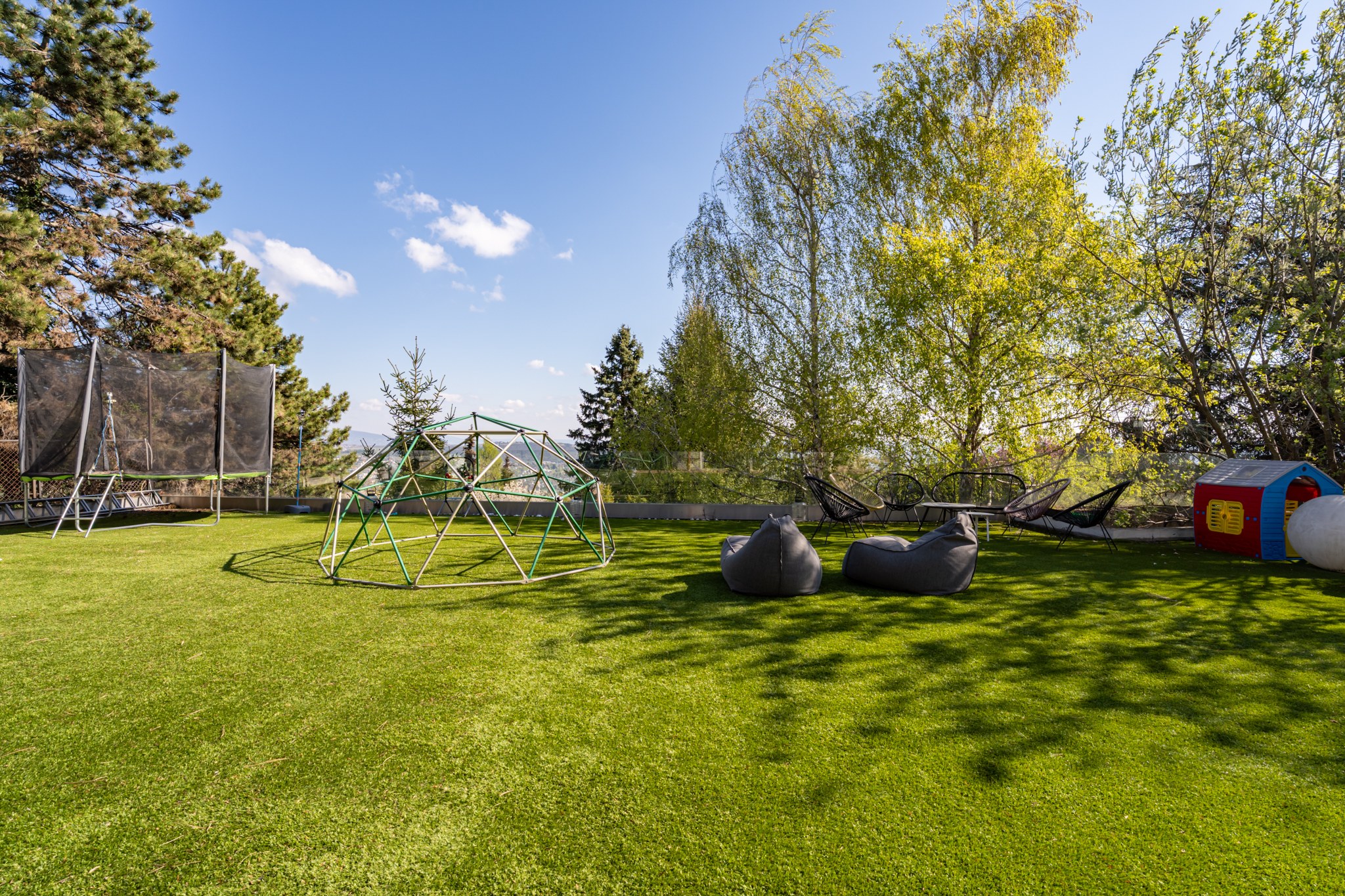 A spacious, sun-drenched garden with artificial grass, play equipment and a relaxation corner. Trees and a distant view complete the backdrop.