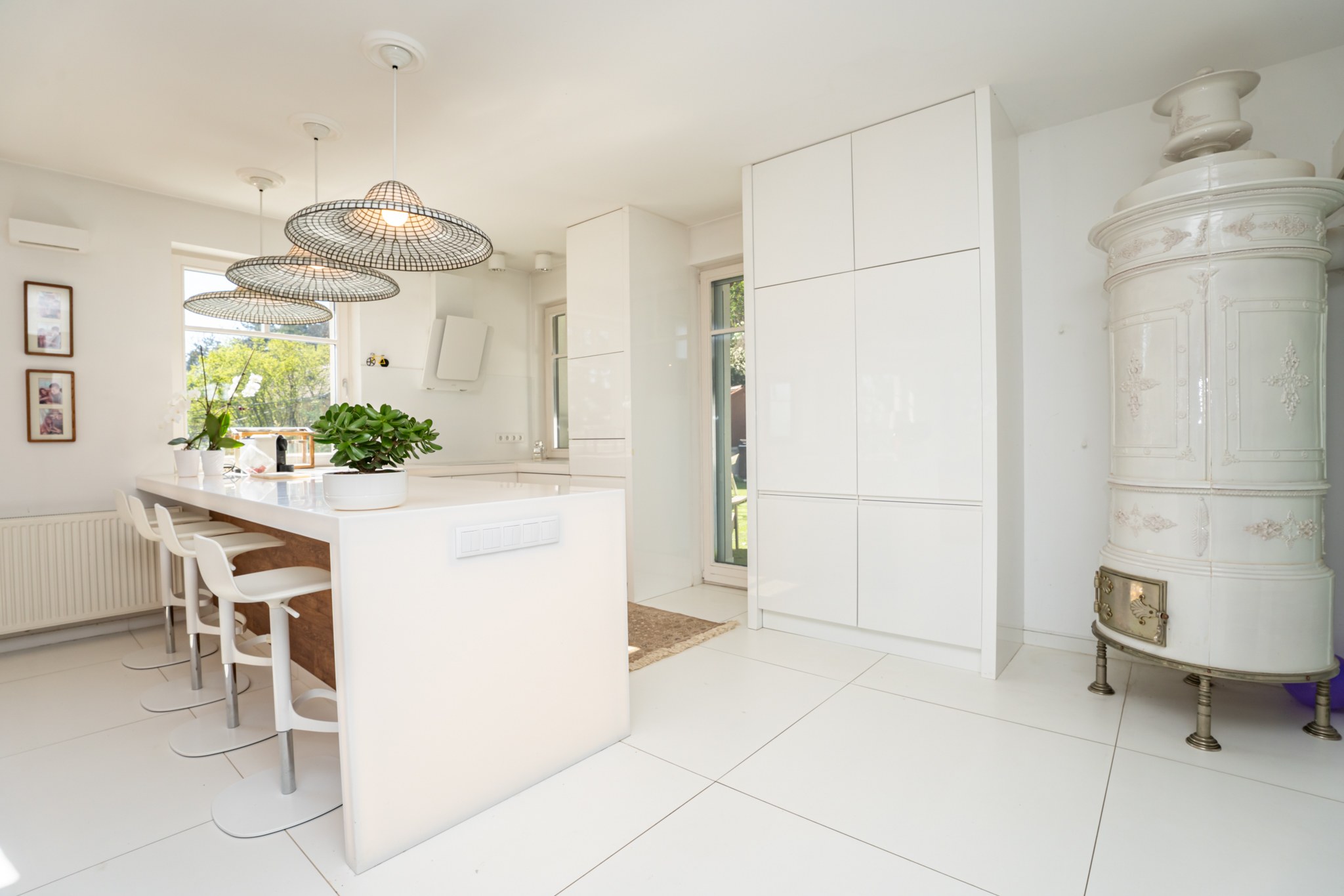 A modern, bright kitchen with white cabinetry and an island with bar stools. A traditional tiled stove also features in the room, while greenery is visible through the windows.