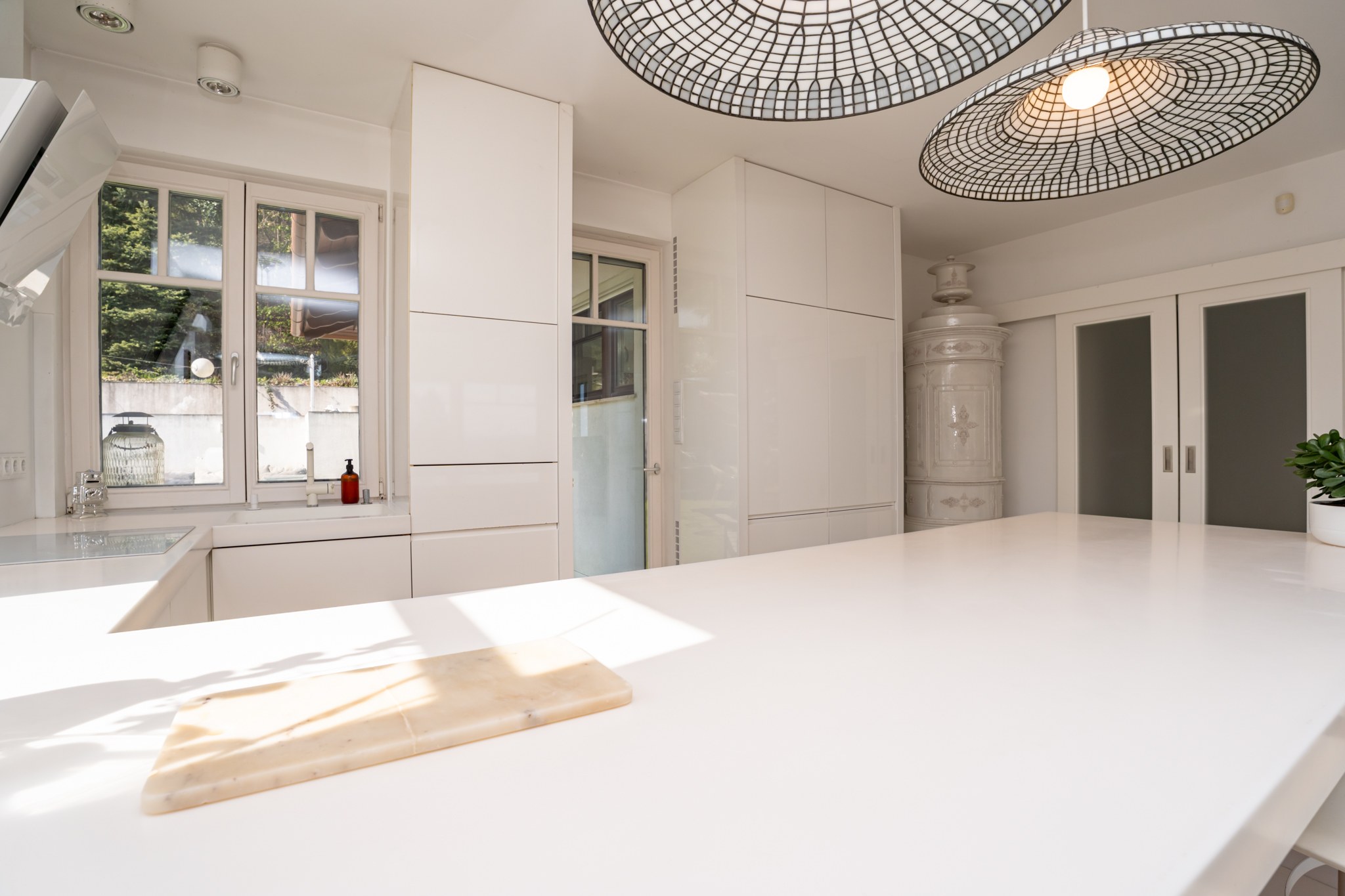 A bright, modern kitchen with a large white island and elegant integrated cabinetry. Abundant natural light streams through the windows, catching the countertop and chopping board. The space is made distinctive by a vintage-style tiled stove and two modern mesh pendant lights. The kitchen is clean, tidy and welcoming.