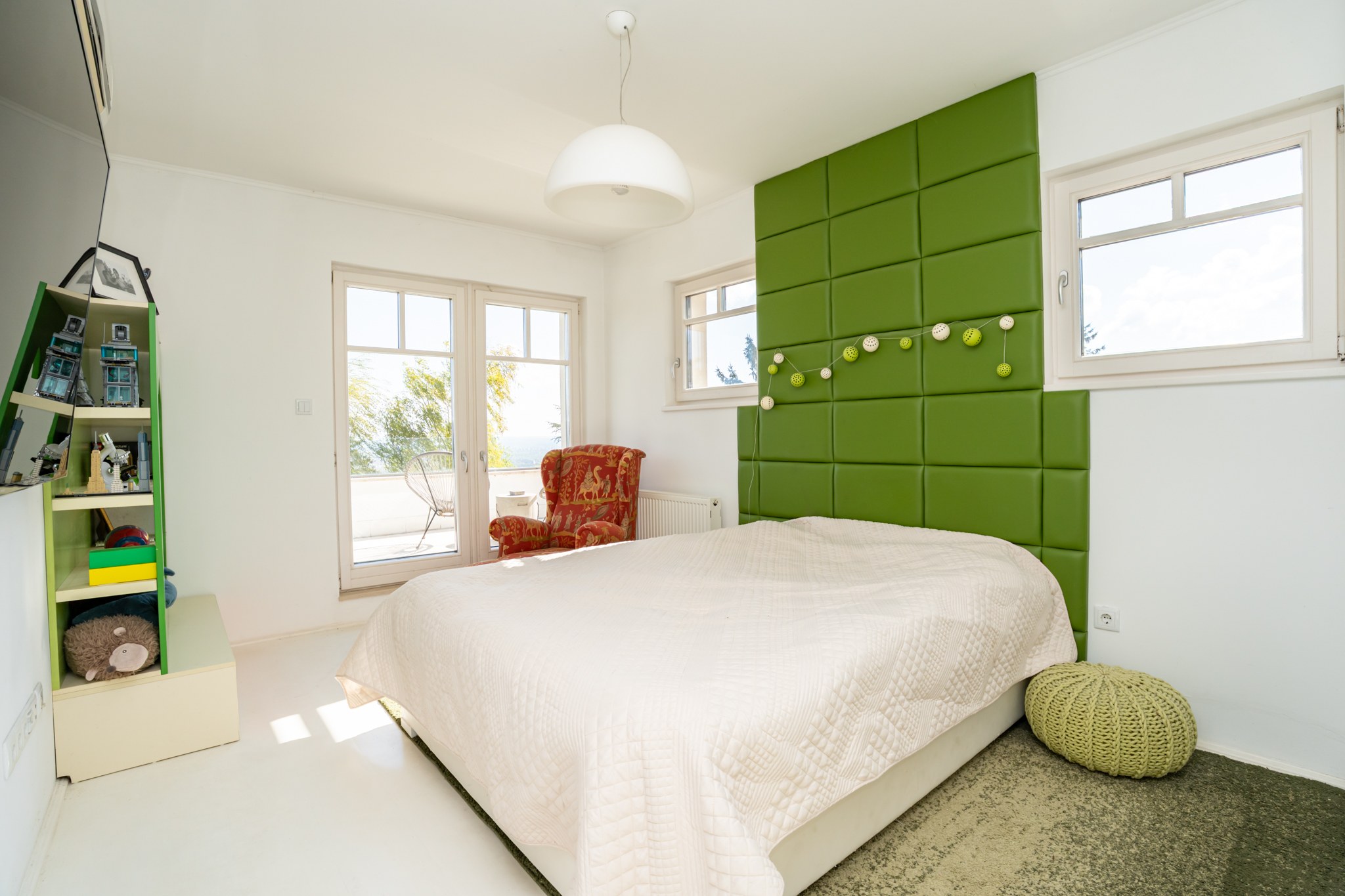 A bright bedroom with modern furnishings and large French doors admitting abundant natural light. The focal point of the room is a generously sized bed with a green upholstered headboard.