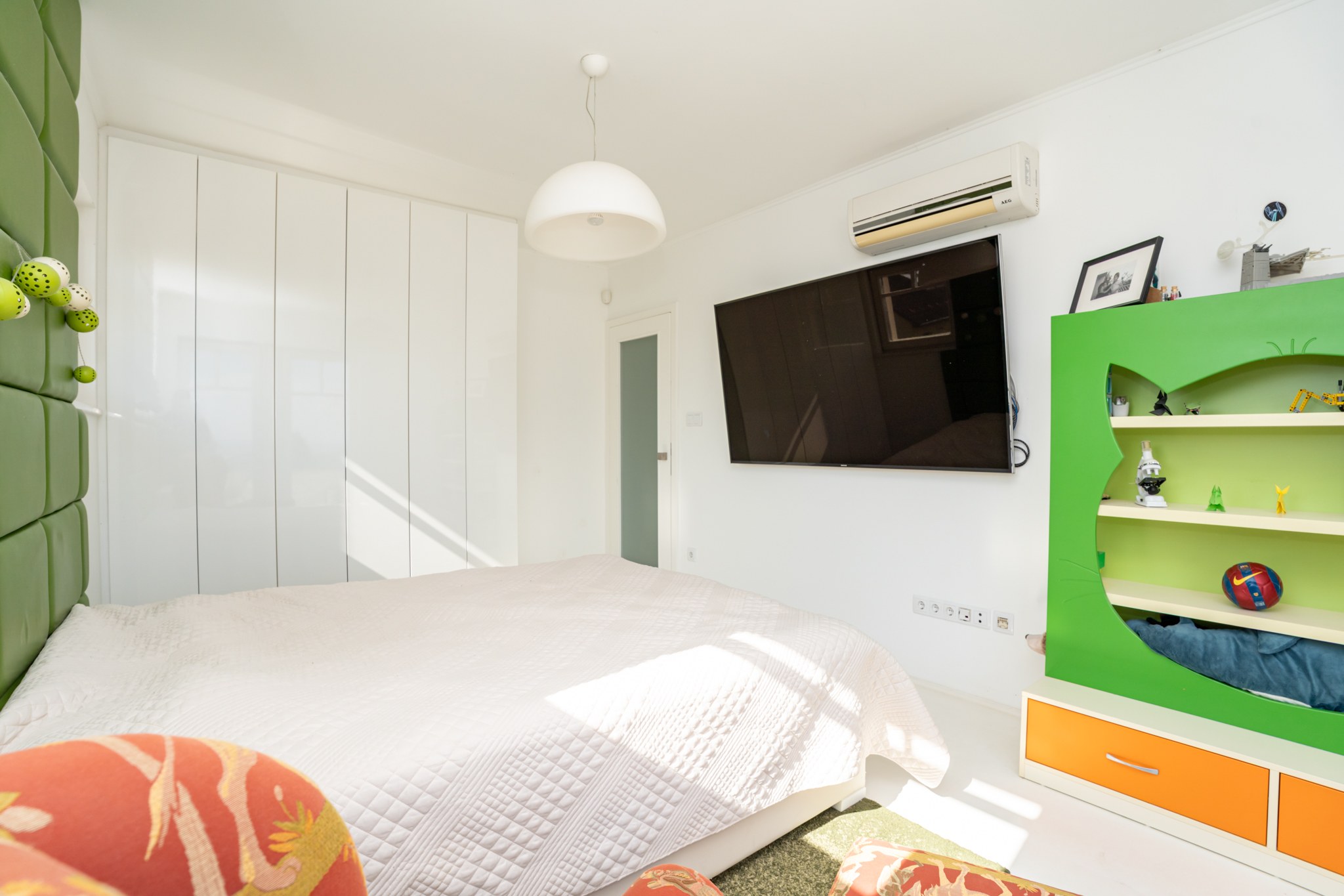 A modern, bright bedroom or children's room where white and green tones predominate. The space is clean and tidy, bathed in sunlight. A wall-mounted TV and air conditioning unit are also part of the room.