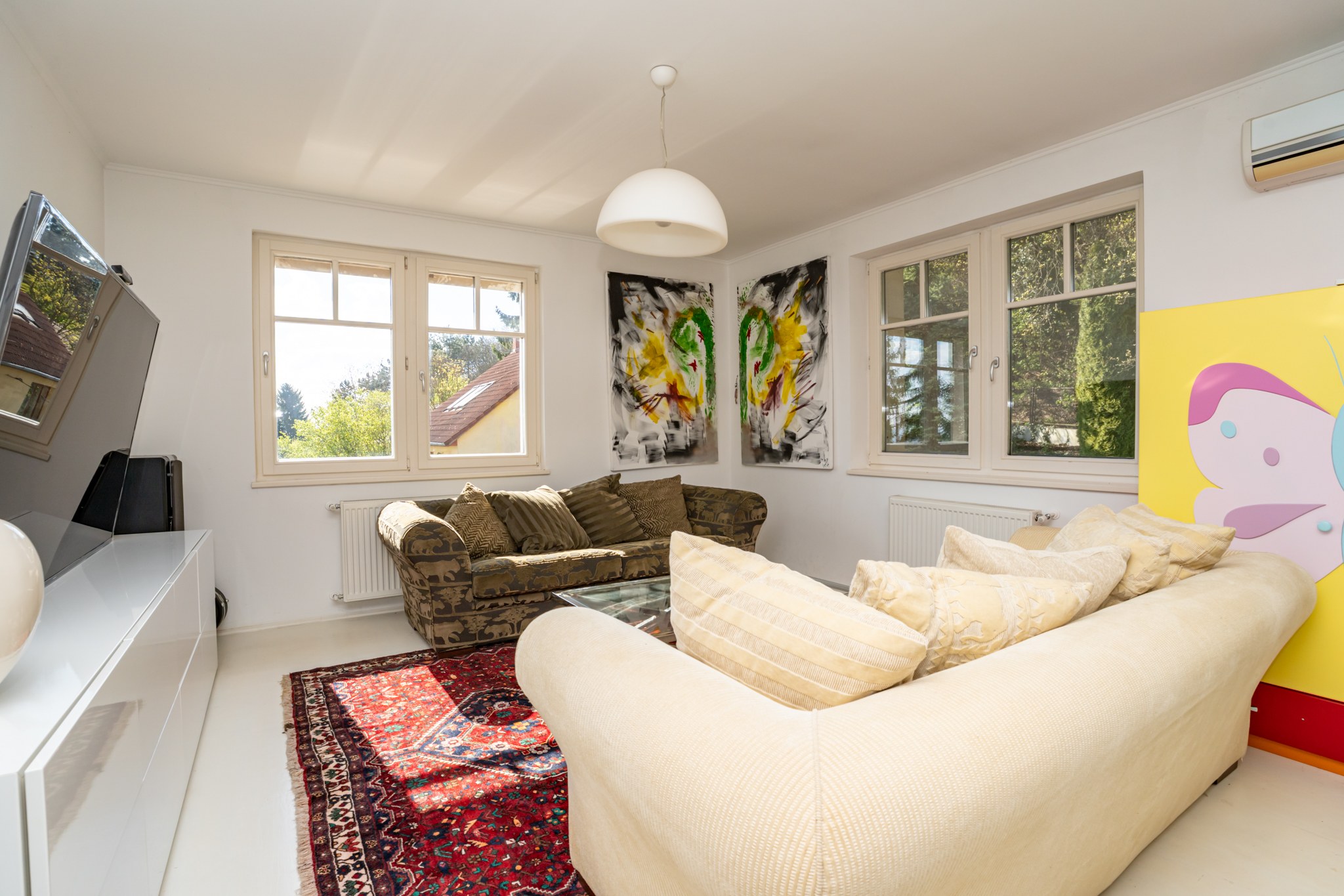 A bright and modern living room with large windows through which abundant natural light pours. The room is furnished with comfortable sofas, modern artworks and a large television, creating a distinctive atmosphere.