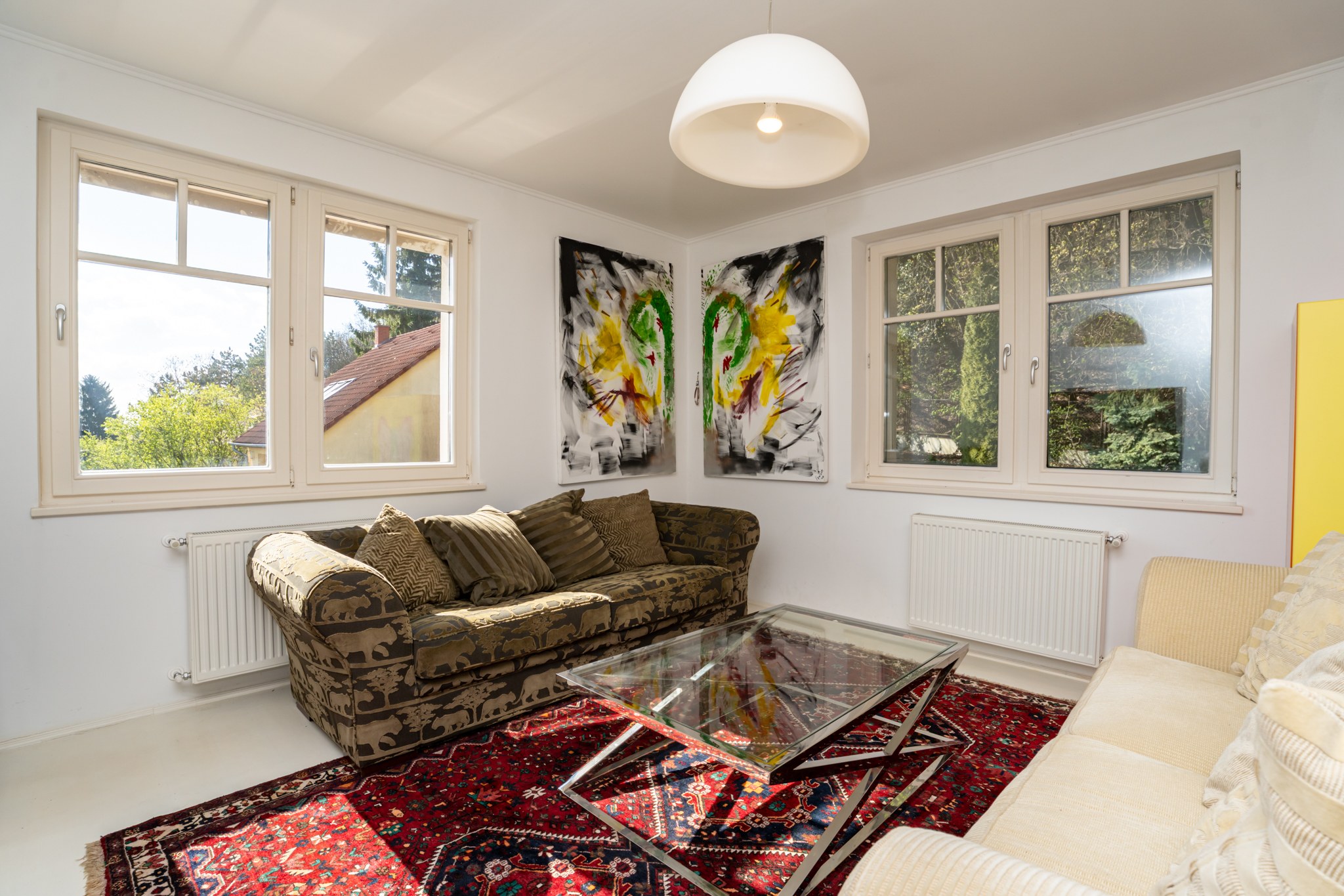 A bright living room with two sofas and a glass coffee table. Large windows admit abundant natural light with views of the surrounding trees and a neighbouring house. The room is decorated with modern and abstract paintings and a patterned Persian rug, creating a warm, homely atmosphere.