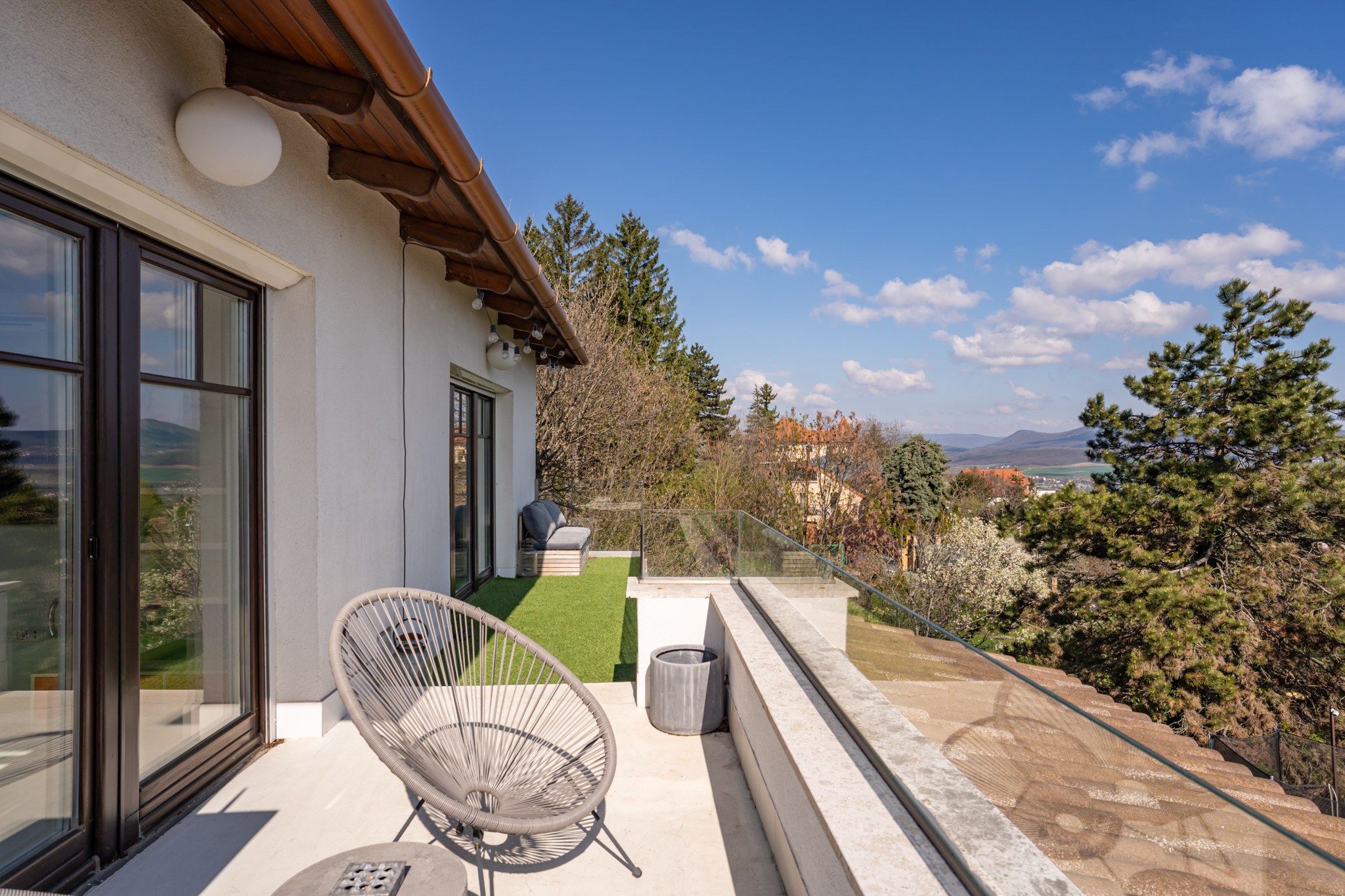 This spacious terrace is furnished with modern garden furniture and artificial grass, offering panoramic views of the landscape and a distant settlement. Sunny and bright, it shows the transitional outdoor space opening from the property towards the garden.