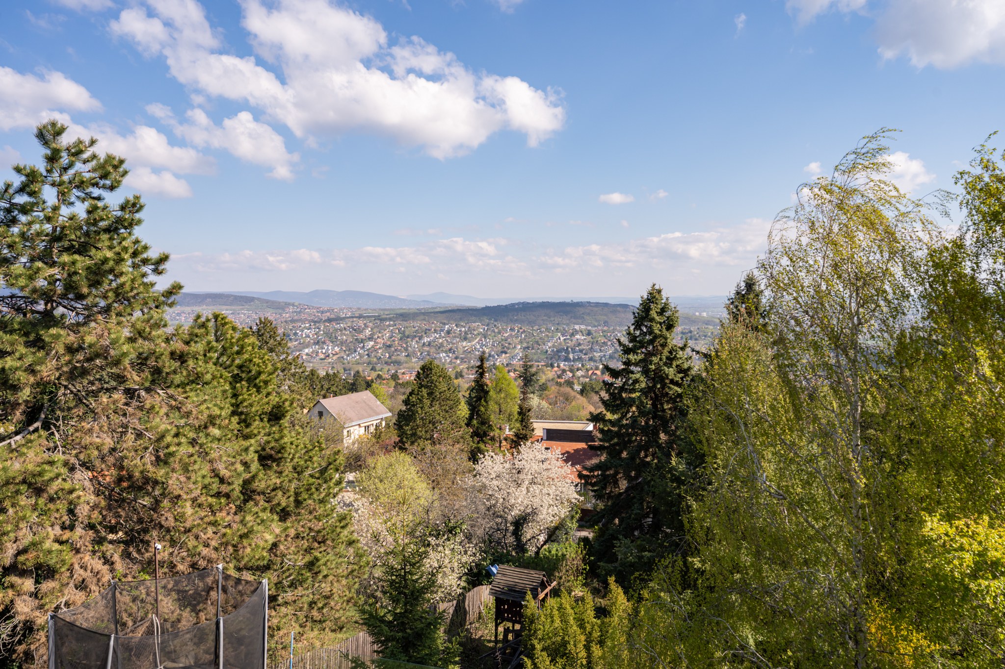 Panoramic view over a densely built residential hillside neighbourhood, with distant mountains and a clear blue sky with white clouds. In the foreground, lush green vegetation, several rooftops and a trampoline are visible.