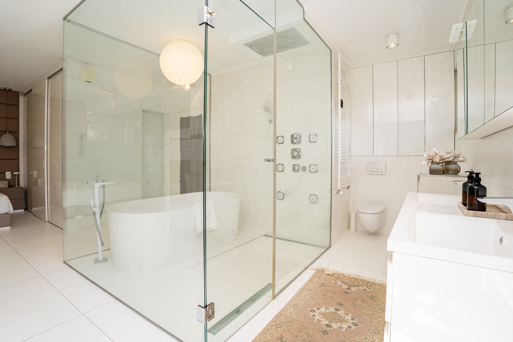 A modern, elegant bathroom connecting to the bedroom via a glass wall. Light, clean white finishes dominate, with quality fittings throughout.