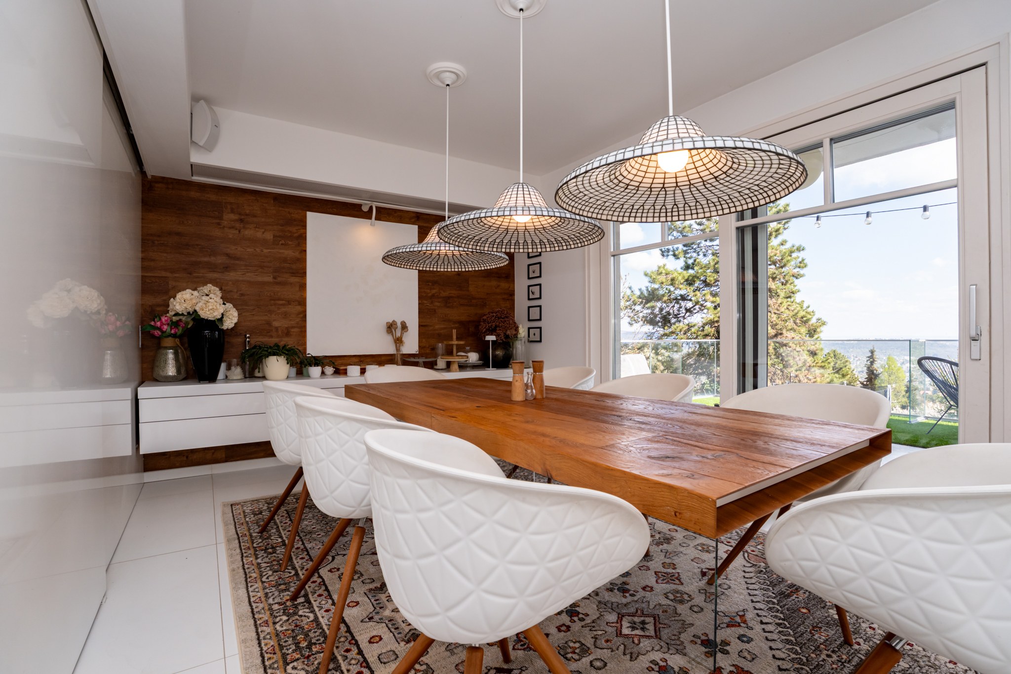 A bright, modern dining room with large windows and panoramic views. A substantial wooden dining table takes centre stage, surrounded by stylish white chairs. Part of the wall is clad in a wood-effect finish.