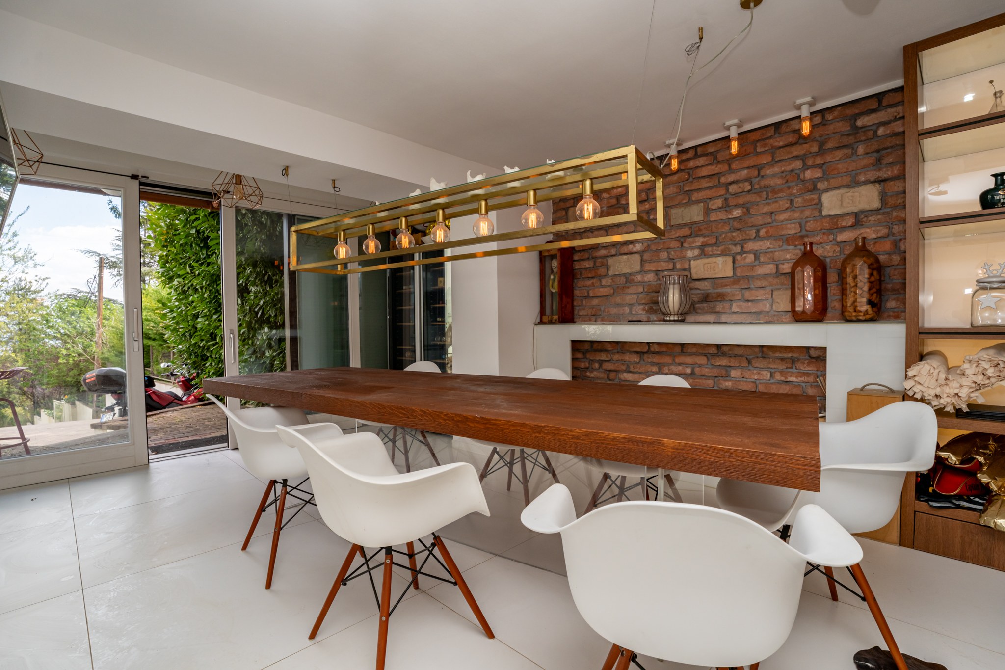 A modern and spacious dining room centred around a large wooden dining table with contemporary white chairs arranged around it. One wall features raw brick cladding, adding character to the space. Large sliding glass doors open onto a green outdoor area, admitting generous natural light.