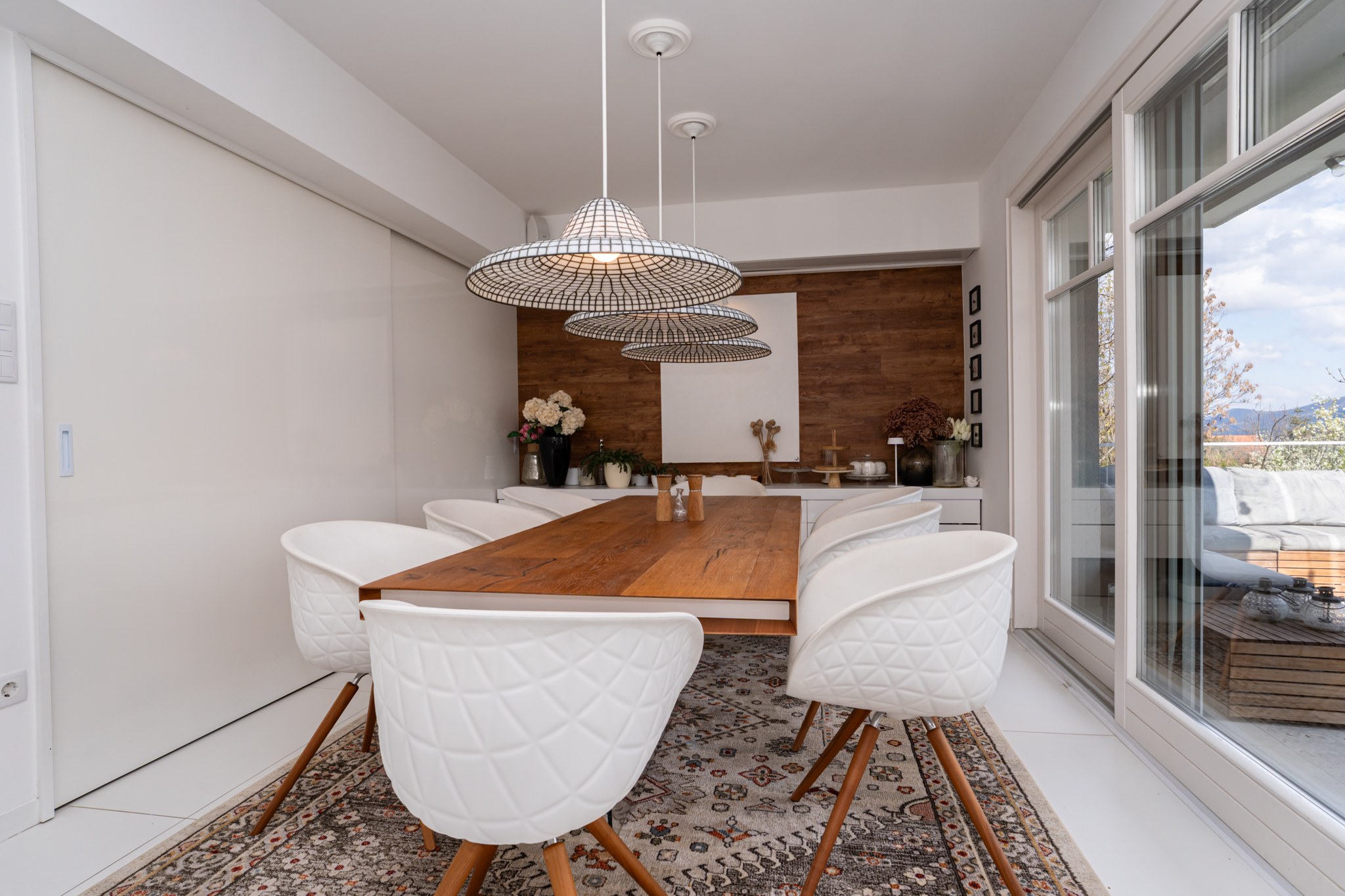 A bright and modern dining room whose sliding door opens onto a spacious terrace with views of the surrounding landscape. The interior is furnished with an elegant wooden table and white patterned chairs, with a wood-clad wall and a floating shelf visible.