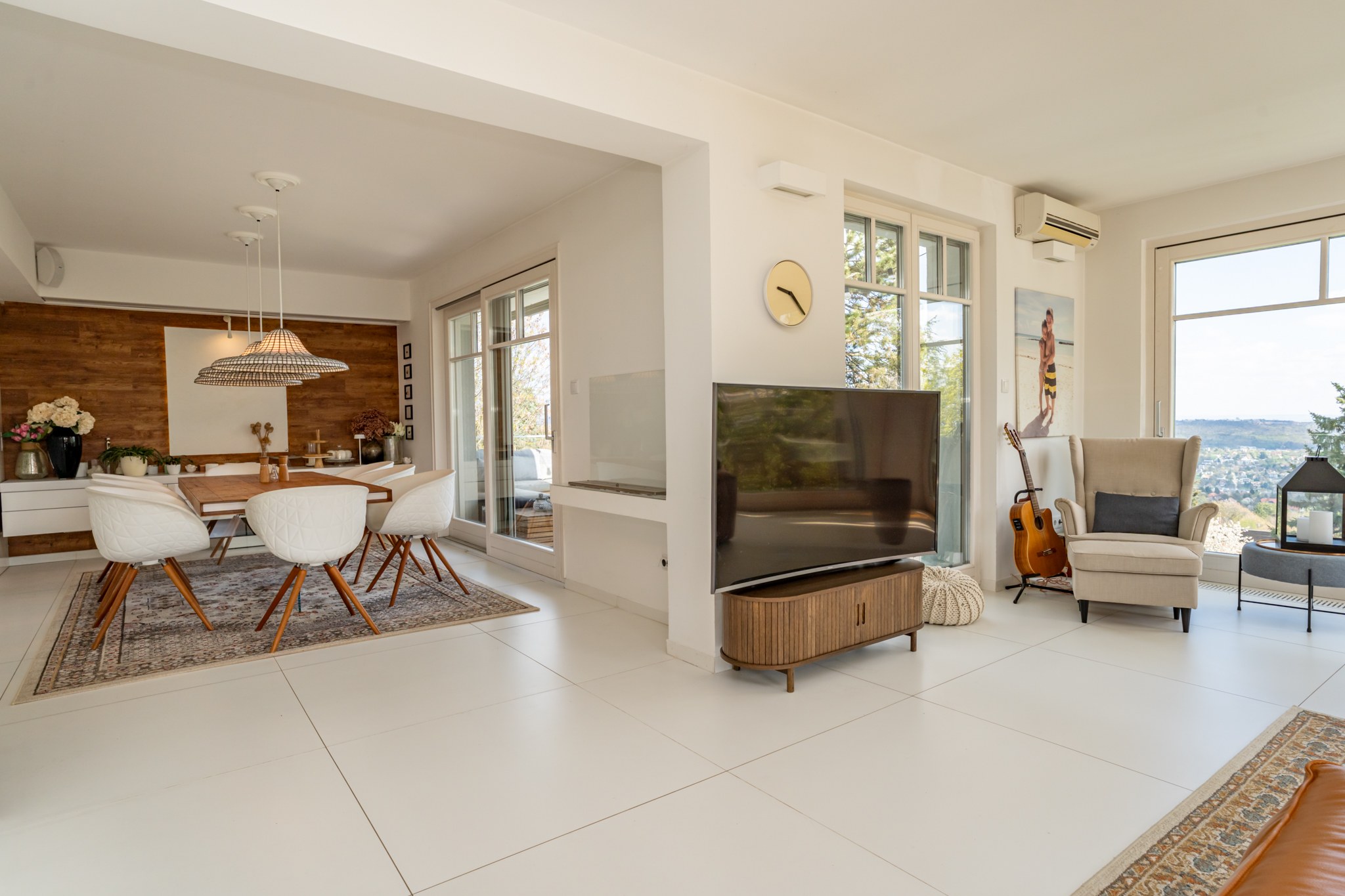A bright, modern open-plan living-dining room with white finishes and wooden decorative elements. Large windows provide abundant natural light and panoramic views of the landscape and cityscape.