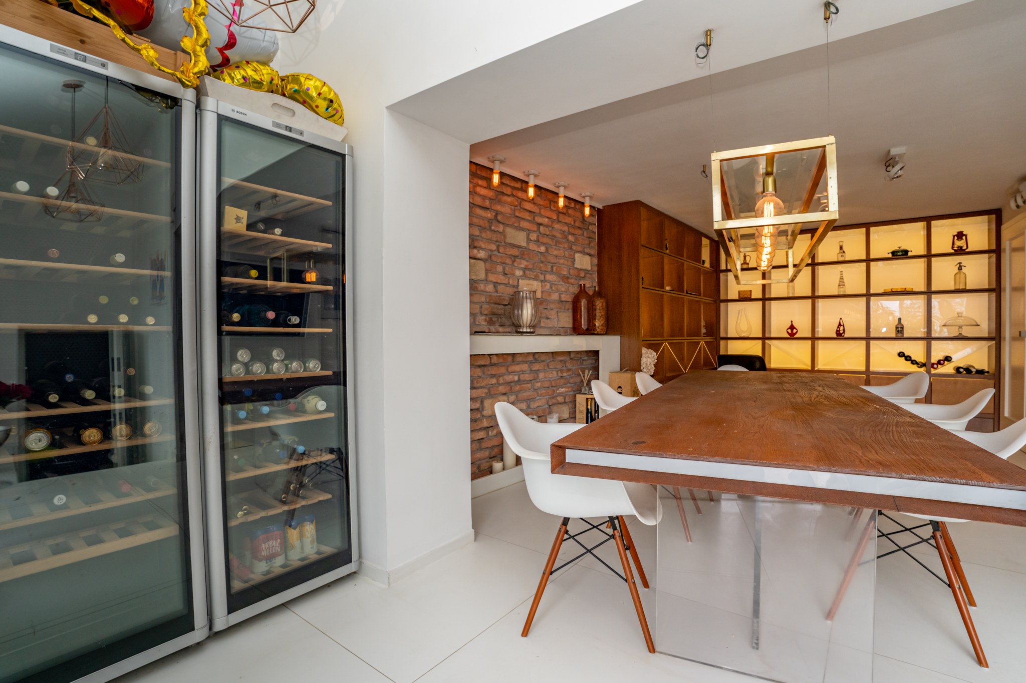 A modern dining room dominated by a large, substantial wooden dining table surrounded by stylish white designer chairs. Two large wine coolers, a brick-clad wall and built-in backlit shelving create an atmospheric setting.
