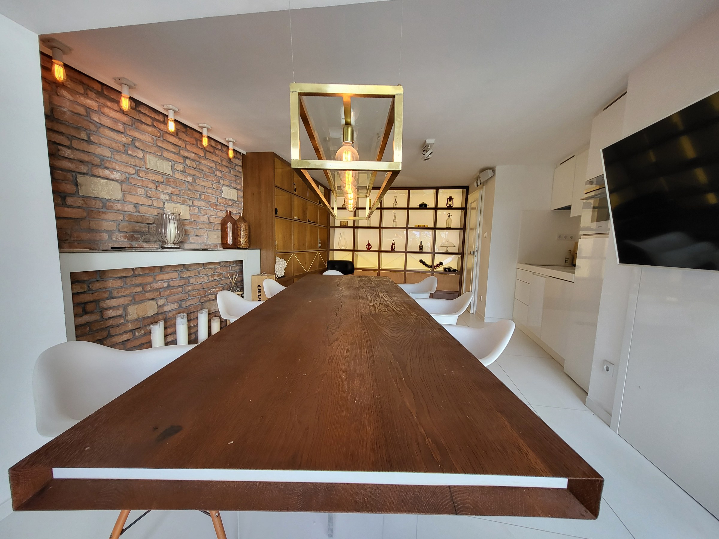 A modern, elongated dining room with a large wooden dining table at its centre. A decorative brick wall runs along one side, highlighted by spotlights, with a built-in shelving unit opposite. A glimpse of the kitchen and a wall-mounted TV appear in the background, lending the space a modern and refined character.